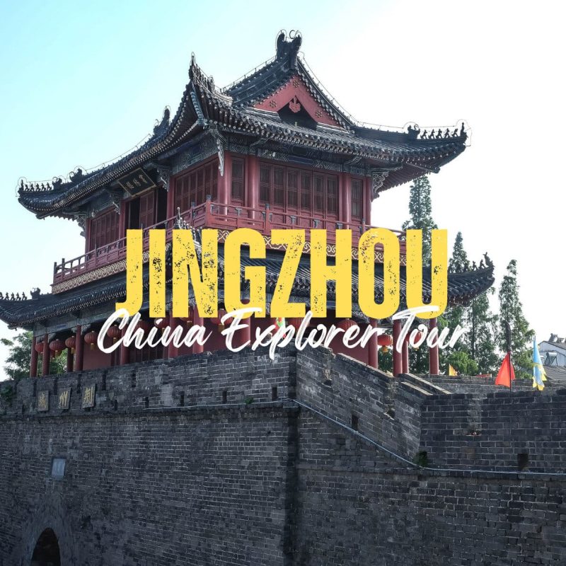 Jingzhou,Hubei,Chinatravel Historic Chinese tower perched on a stone city wall, with large yellow text 'JINGZHOU' and cursive 'China Explorer Tour' overlay against a blue sky.