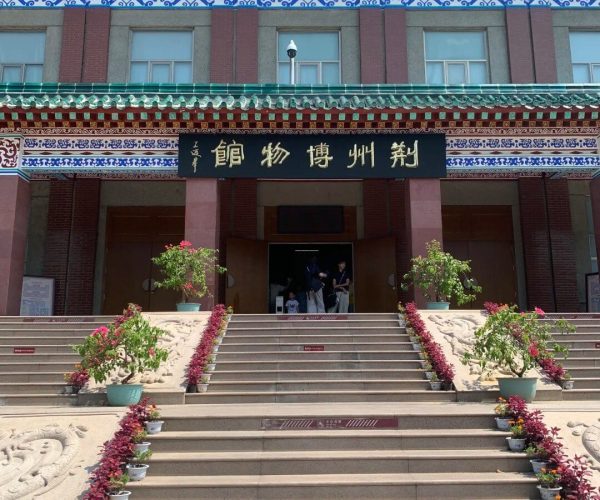 Jingzhou Museum,Jingzhou,Chinatravel Front entrance of a Chinese museum with ornate blue and red decorative friezes, green-tiled roof, and a large black sign with gold Chinese characters above the doors; steps lined with potted plants lead up to the entrance.
