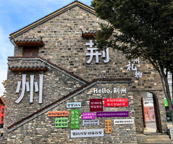 Shashi Old Port,Jingzhou,Chinatravel Brick building with large white Chinese characters and the words 'Hello, 荆州' on its wall, colorful signs pasted across the lower facade, and a tree partially obscuring the right side.