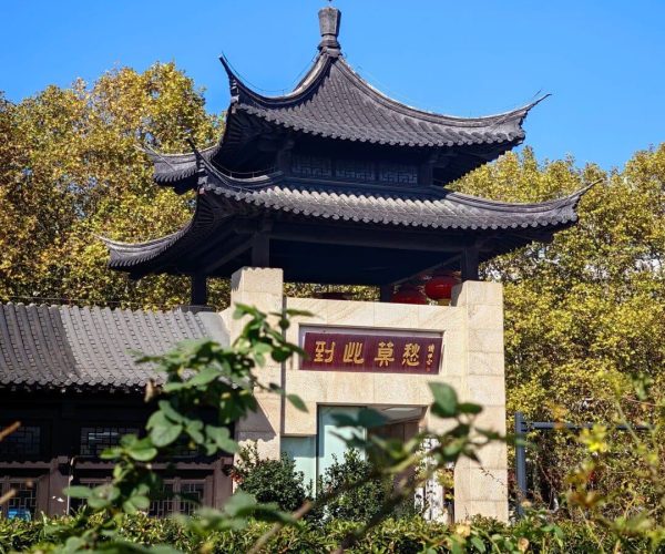 Mochou Lake Park,Nanjing,China tour Traditional Chinese pavilion atop a stone gate with red lanterns and Chinese signage, framed by green bushes and a clear blue sky.