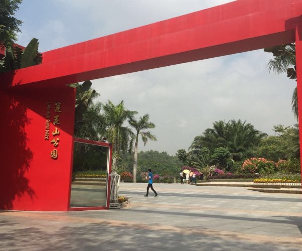 Lianhua Mountain Park,Shenzhen,Chinatravel Bright red gateway arch marks a park entrance, with Chinese characters on the left pillar and tropical plants beyond.