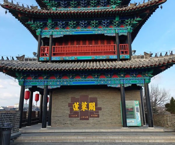 Penglai Pavilion,Yantai,China tour Colorful Chinese pagoda-style gate with two tiers of upturned roofs, red lattice panels, and lanterns against a blue sky on a brick wall site wall.