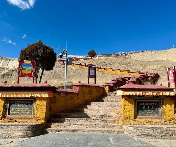 Tomb of the Tibetan King,Shannan,China tour Stone steps ascend a hillside flanked by yellow-red stone walls, with prayer flags along the slope and several signboards at the entrance under a clear blue sky.