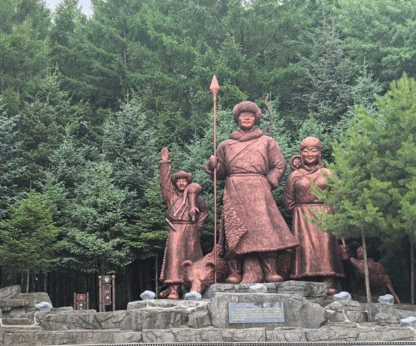 Jiejinkou Hezhen Ethnic Customs Park,Jiamusi,Chinatravel Bronze statue group of three people in traditional clothing with a spear, on a stone pedestal in a pine forest park.