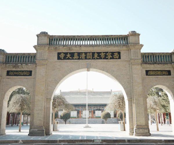 Dongguan Mosque,Xining,Chinatrip Stone city/temple entrance arch made of beige brick, with a central black plaque featuring gold Chinese characters above the arch; courtyard beyond with trees and a long building visible in distance.