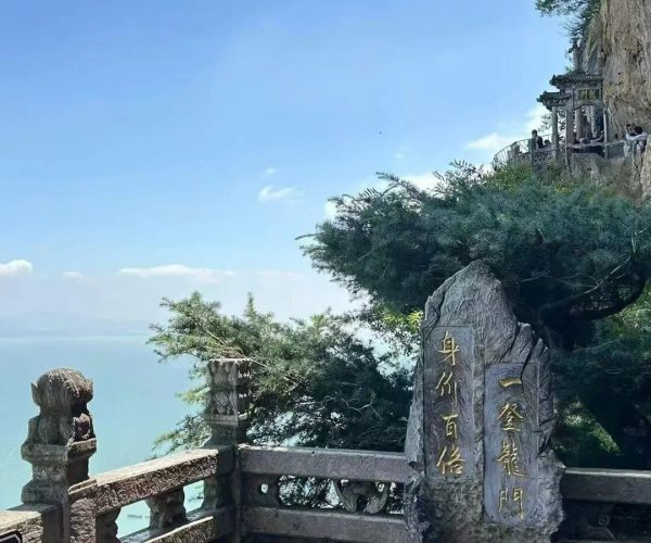 Western Hills & Dragon Gate,Kunming,Chinatravel Cliffside lookout with stone railing, golden Chinese inscriptions on rocks, and visitors along a mountain path against a blue sky and distant sea.