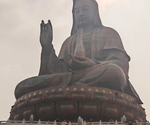 Xiqiao Mountain,Foshan,Chinatravel Large seated Buddha statue on a wide pedestal, one hand raised in blessing and the other holding a vase, against a pale sky.