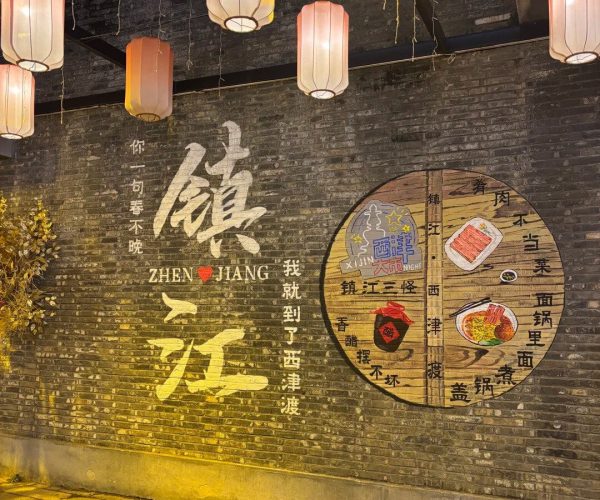 Xijin Ferry,Zhenjiang,China tour Night scene of a brick wall mural with large Chinese characters and a round diagram, illuminated by pink lanterns overhead and warm street lighting.