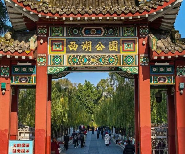 West Lake Park,Fuzhou,Chinatravel Ornate Chinese gate with red columns and dragon decorations opens to a tree-lined park; people walk along the path under a bright blue sky.