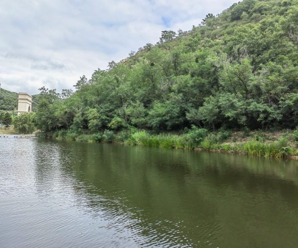 Xijiao Lake Wetland Park,Jingzhou,Chinatravel Calm river runs beside a forested bank, with a cream stone arch structure visible on the left and green hills in the background.