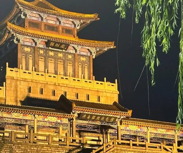 Liberation Pavilion,Jinan,China tour Night-time view of a lit traditional Chinese pagoda-style tower on a stone wall, with a red star on top and willow branches hanging nearby.