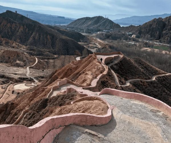 Guide National Geological Park,Hainan Prefecture,China tour Winding pinkish path along rugged hills with a lone person walking, landscape of eroded red clay hills in the distance