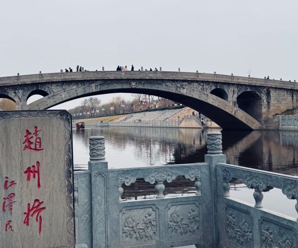 Zhaozhou Bridge,Shijiazhuang,China tour Stone arch bridge crossing a calm river, with pedestrians along the top; decorative railings and a carved stone plaque with red Chinese characters in the foreground.