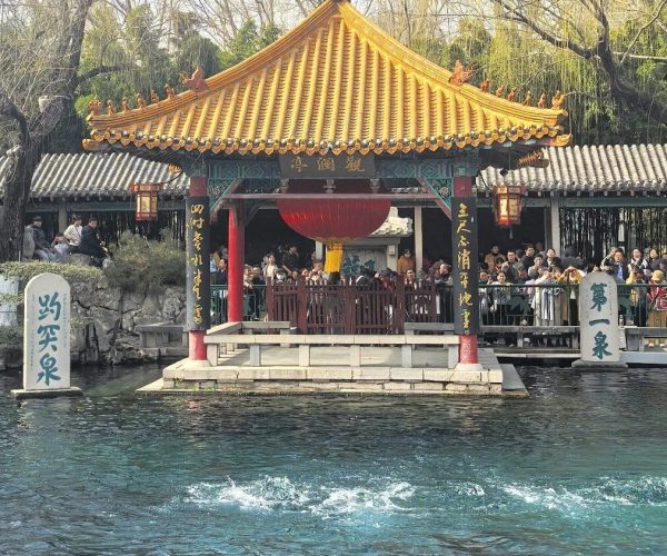 Baotu Spring,Jinan,China tour Traditional Chinese pavilion with a yellow tiled roof on a water-filled terrace, red columns, and a crowd nearby by a canal.