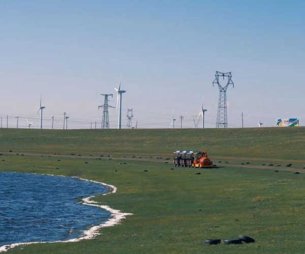 Huitengxile Grassland,Hohhot,Chinatravel Wide coastal plain with a blue lake on the left, wind turbines and transmission towers on the horizon, and an orange maintenance vehicle with passengers near the center.