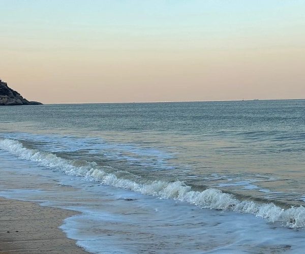 Lian Island,Lianyungang,China tour Calm sea at a sandy beach with gentle waves; a rocky headland with buildings on the left under a pastel sky.