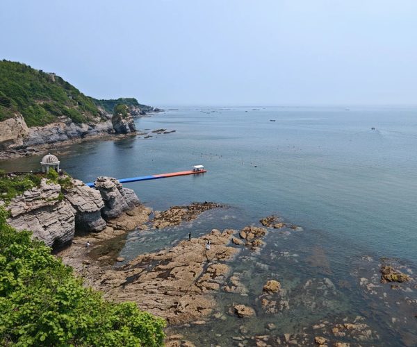 Golden Pebble Beach (Jinshitan) National Geopark,Dalian,China tour Coastal rocky shoreline with a small domed pavilion on a cliff, a colorful floating platform extending into calm blue water, and people exploring the rocks below.
