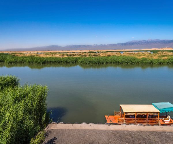 Yinchuan Weather,Yinchuan,Chinatrip Calm river with reed-lined banks, desert dunes and distant mountains under a blue sky; a wooden canopy boat is docked at the shore.