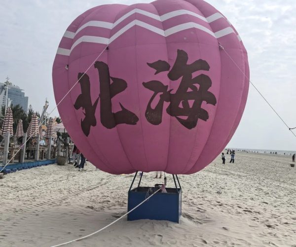 Silver Beach,Beihai,Chinatravel Pink hot air balloon with black Chinese characters tethered to a blue base on a sandy beach, with people and buildings in the background.
