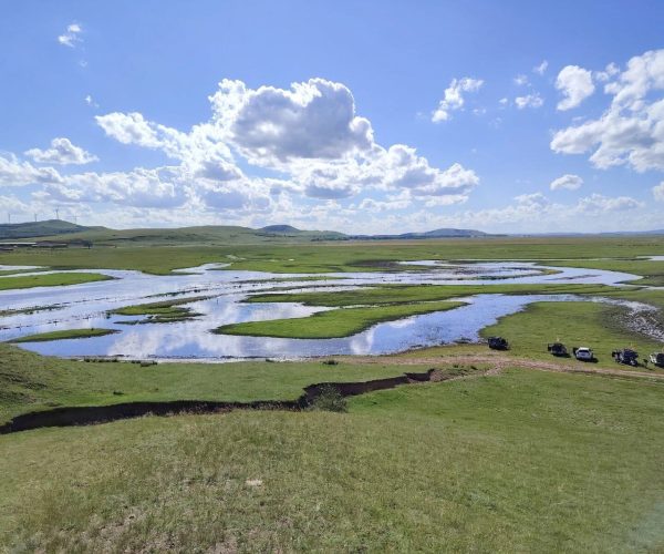 Huitengxile Grassland,Hohot,Chinatrip Wide grassland with a winding river reflecting the blue sky; distant hills and a line of parked cars on the right side of the image.
