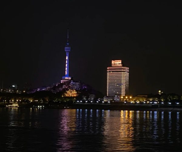 Wuhan Yangtze River Cruise,Wuhan,Chinatravel Night cityscape featuring a tall illuminated tower atop a hill and a striped lit hotel by the river.