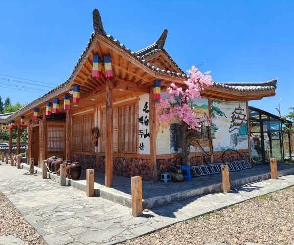 Changbai Korean Folk Village,Baishan,Chinatrip Traditional Korean hanok-style building with wooden beams, colorful lanterns, and a stone-paved path under a bright blue sky, pink blossoms nearby