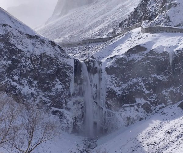 Changbai Waterfall,Baishan,Chinatrip Snowy mountain landscape with a tall waterfall pouring down a rocky cliff, a long curved stone wall snaking along the ridge, and leafless trees in the foreground.