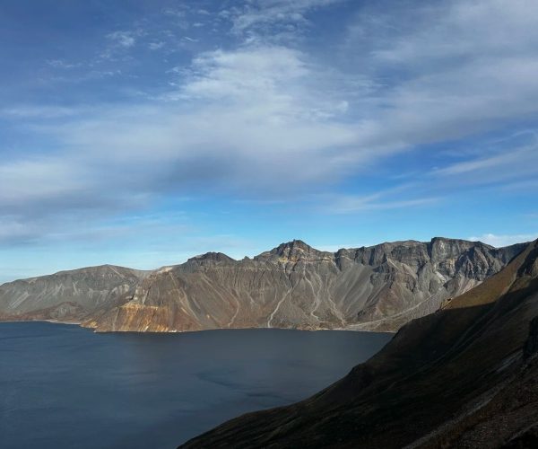 Baishan West Slope Scenic Area,Baishan,Chinatrip Wide view of a dark blue crater lake edged by steep brown-gray volcanic cliffs and jagged mountains under a blue, partly cloudy sky.