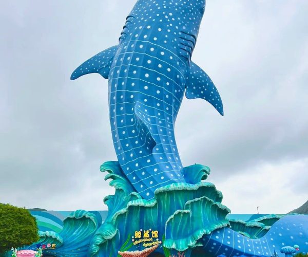 Chimelong Ocean Kingdom,Zhuhai,Chinatravel Giant blue whale shark sculpture with white spots rising above turquoise waves, at a theme park entrance with visitors nearby.