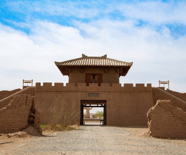 Yangguan Ancient Pass,Dunhuang,Chinatrip Mud-brick fortress gate with a tiled roof tower, crenellated walls, and a desert landscape beneath a blue sky.