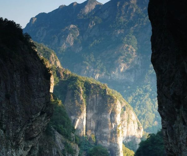 Yandang Mountain,Wenzhou,Chinatravel View of a narrow canyon with towering rocky cliffs and forested slopes under a clear blue sky, a winding road at the bottom left edge.