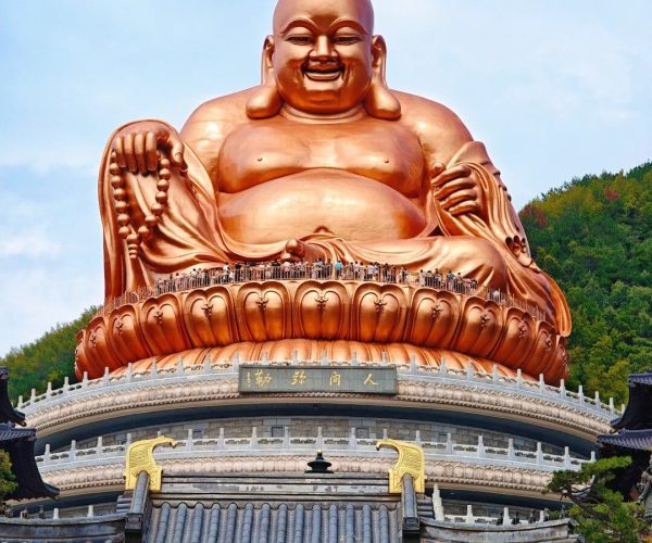 Xuedou Temple,Zhejiang,Chinatravel Gigantic golden Laughing Buddha statue seated on a lotus pedestal above a traditional temple roof, with visitors at the base under a blue sky.
