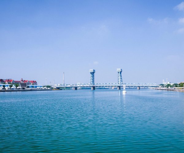 QingdaoWeather,Qingdao,Chinatravel Blue sky over a calm river, with a metal lift bridge spanning the water and tall white buildings along the right shore.