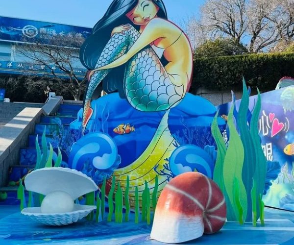 Qingdao Underwater World,Qingdao,Chinatravel Colorful mermaid sculpture with long blue hair seated on stylized waves, surrounded by seashells and seaweed in an ocean-themed display.