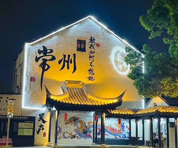 Qingguo Lane,Changzhou,China tour Night view of a traditional Chinese building with a lit roofline and large calligraphy on the wall; dragon mural along the lower wall. Interpretive signage suggests a cultural site or theater.