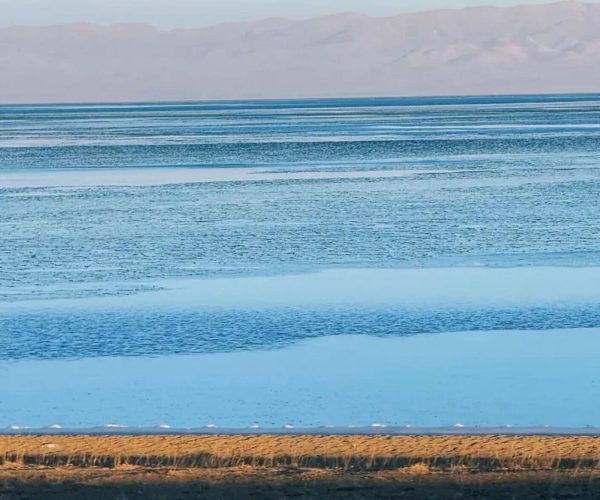 Qinghai Weather,Qinghai,China tour Calm seaside scene: blue water with gentle waves and a sandy shore in the foreground, distant mountains on the horizon.
