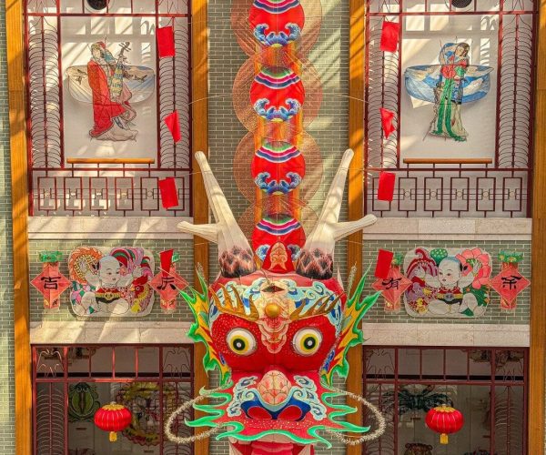 Weifang Kite Museum,Weifang,China tour Colorful Chinese dragon head lantern with green highlights, hanging red tassels, in front of a tiled temple wall with festive red decorations and murals