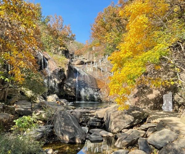 Yuwan Scenic Area,Lianyungang,China tour Autumn scene of a rocky creek with a small waterfall, gold and orange leaves, and a clear blue sky.