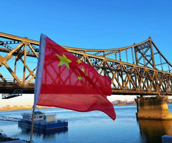 Yalu River Broken Bridge,Dandong,China tour Red Chinese flag waving in front of a large rust-colored steel truss bridge over a calm river, blue sky above.
