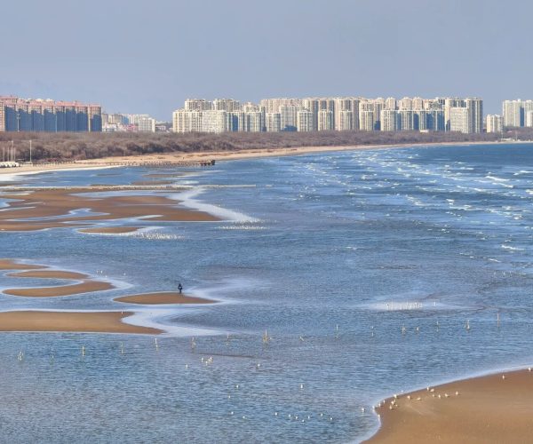 Pigeon Nest Park,Qinhuangdao,Chinatravel Urban skyline along a sandy beach with tidal pools and winding sandbars; a person walks on a sandbar in the foreground.