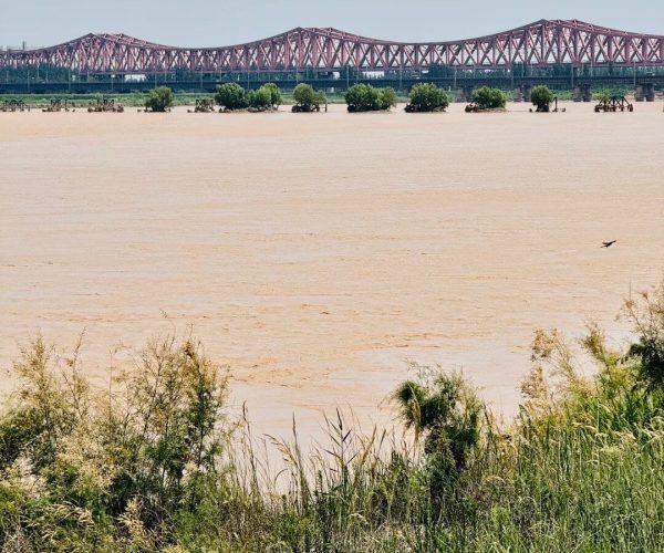 Yellow River Scenic Area,Zhengzhou,Chinatravel Red truss bridge spans a wide muddy river, with grassy riverbank in the foreground and a clear blue sky above.