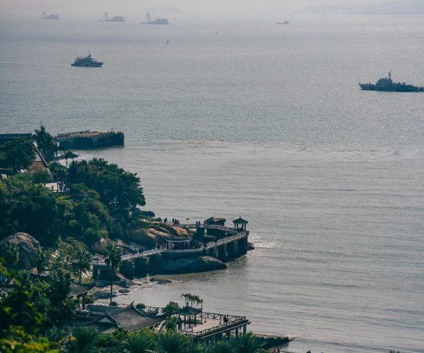 Gulangyu Island,Xiamen,Chinatravel Coastal park with palm trees and a rocky pier, people on the beach, and ships on a calm sea far offshore.