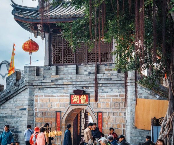 Longhu Ancient Village ,Chaozhou,Chinatravel Stone temple gateway with red lanterns and visitors gathered in a paved square beneath a large tree with hanging roots/branches.