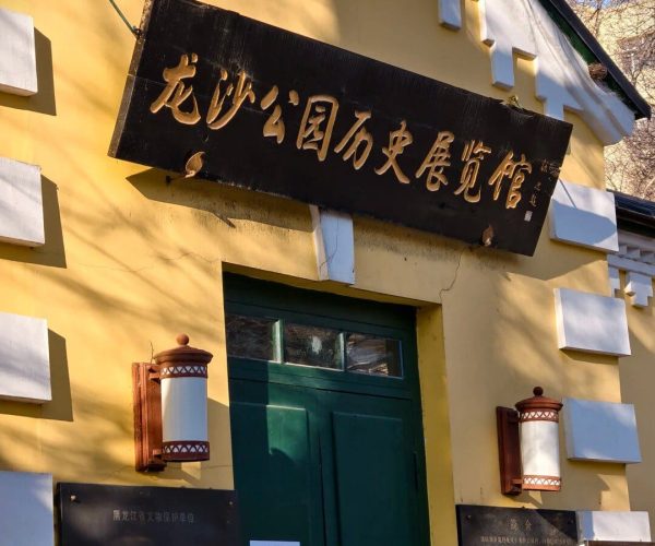Longsha Wetland Park,Qiqihar,Chinatravel Yellow building with a dark wooden sign bearing Chinese characters above a green door, flanked by decorative white blocks and lanterns.