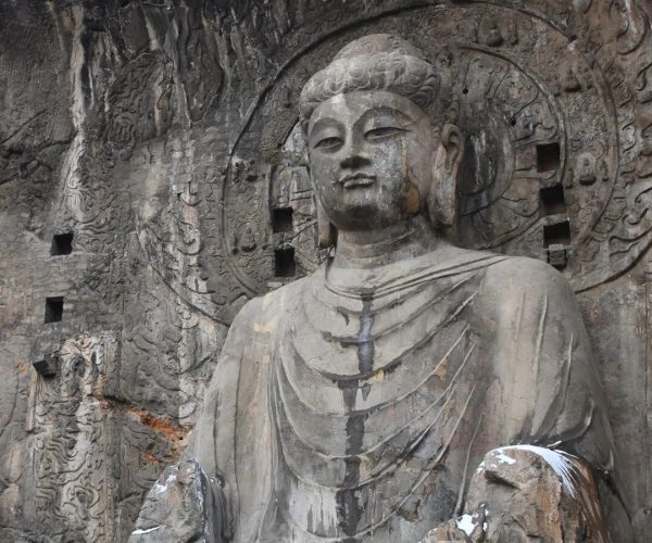 Longmen Grottoes,Luoyang,Chinatravel Rock-cut Buddha statue carved into a cliff with a circular halo behind the head and intricately carved reliefs on the cave wall, with patches of snow on the rock.