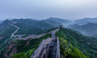 Great Wall segments winding across green mountains under a hazy blue sky with distant ridges.
