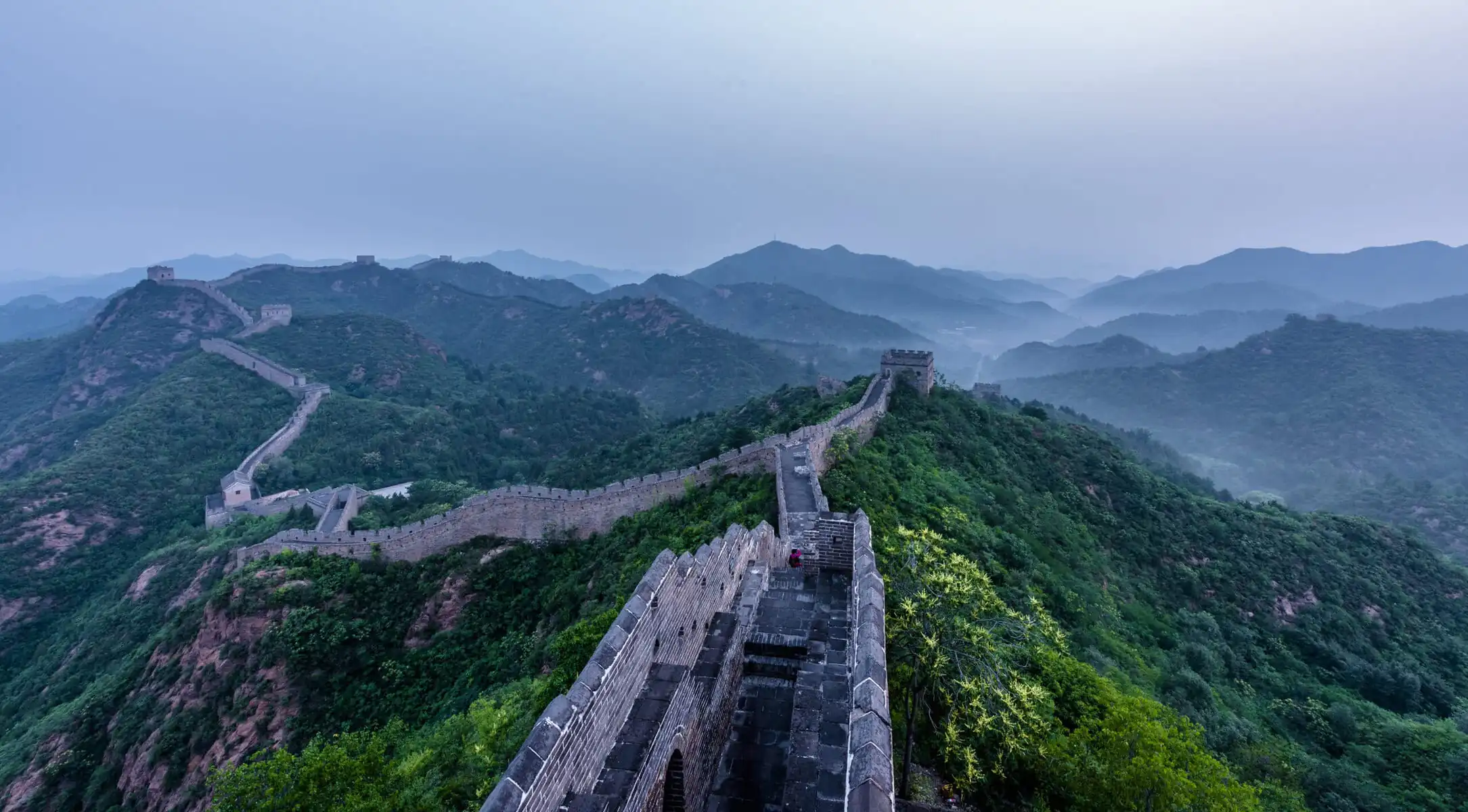 the great wall, Beijing, chinatravel, Great Wall segments winding across green mountains under a hazy blue sky with distant ridges.