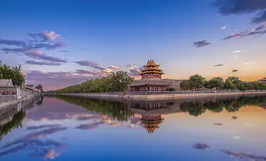 Traditional Chinese pagoda beside a calm moat at sunset, with trees and a stone wall along the bank and visitors on the left.