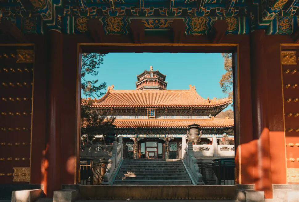 Open gate framing a traditional Chinese temple courtyard, with red columns, ornate gold studs, and a multi-tiered pagoda-style roof against a clear blue sky.