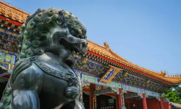 Forbidden City, Beijing, Chinatravel, Bronze guardian lion statue in front of a traditional Chinese temple with ornate, colorful roof tiles against a clear blue sky.